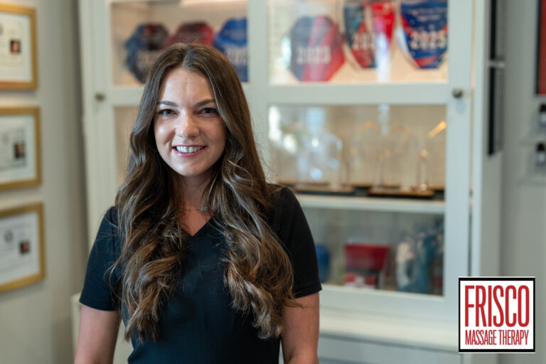 A woman with long brown hair stands smiling in an office with award plaques in the background. The sign in the corner reads "Frisco Massage Therapy," known for therapeutic massage for jaw pain.