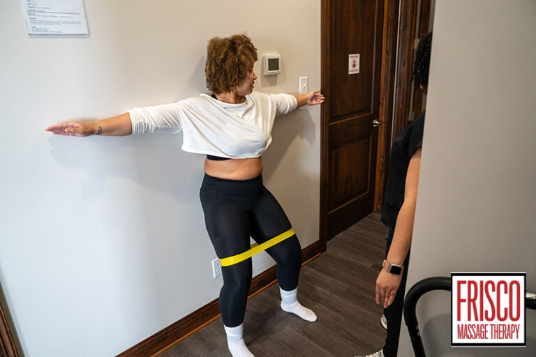 A woman performs a wall exercise with a yellow resistance band around her legs as a therapist observes, showcasing how sports massage for injury prevention is integrated at Frisco Massage Therapy.
