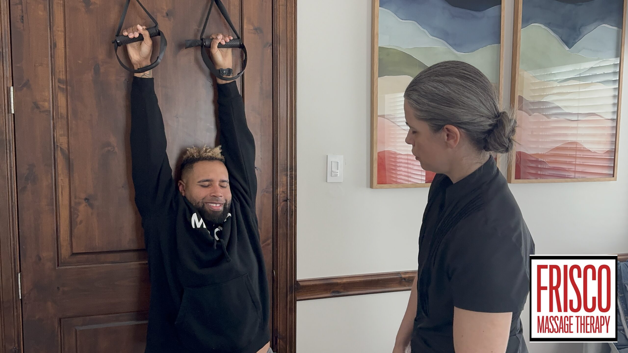 A man hangs from wall-mounted straps while a woman observes, highlighting sports massage for injury prevention; the Frisco Massage Therapy logo appears in the lower right corner.