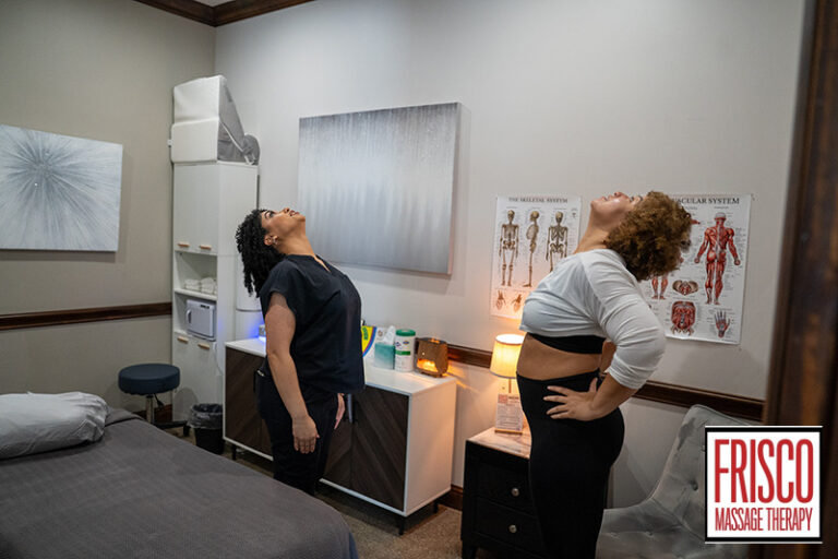 Two women stand in a massage therapy room, tilting their heads back with hands on hips as they perform a neck stretch. Anatomical posters, massage equipment, and cupping supplies are visible in the background.