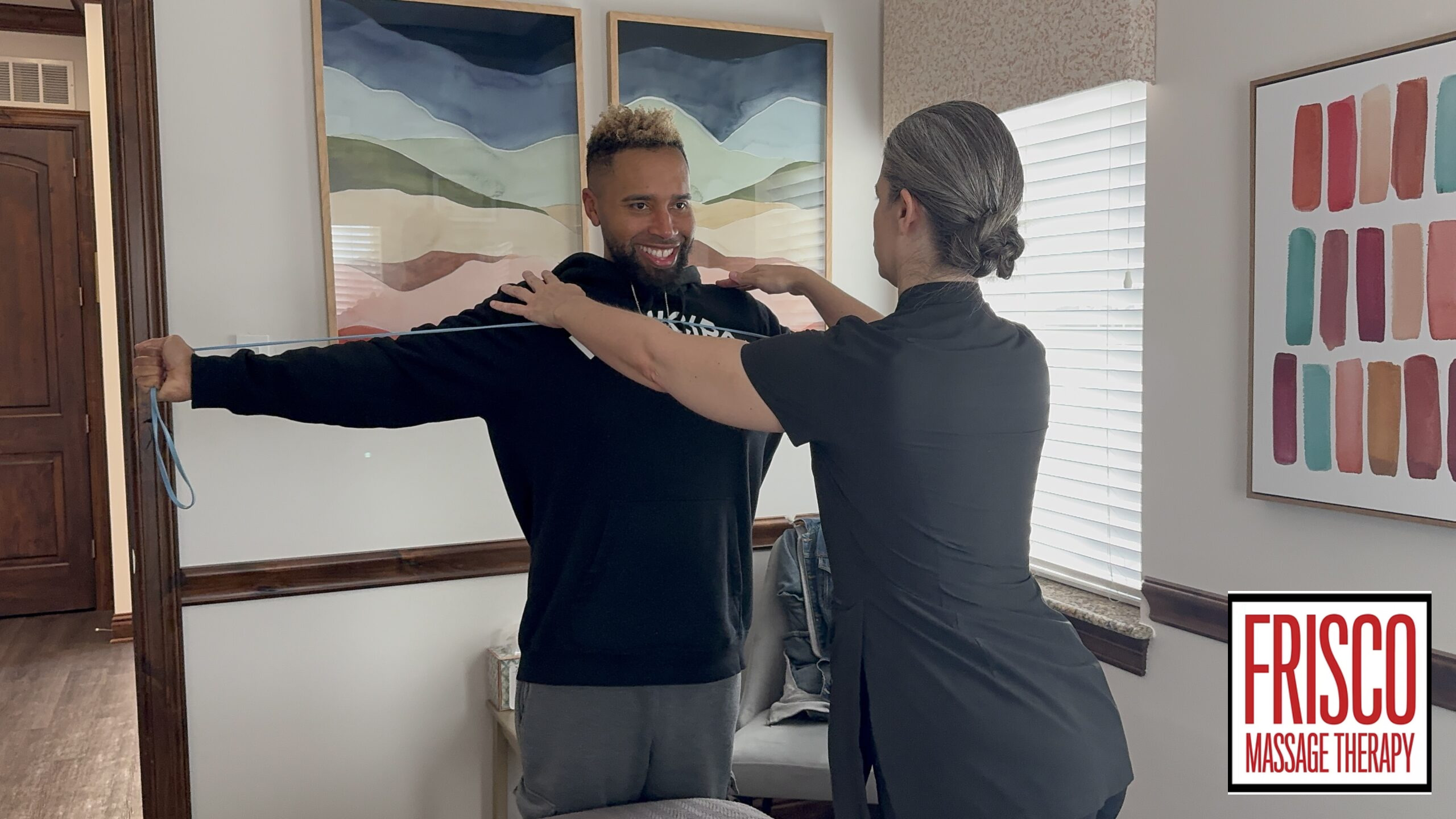 A massage therapist assists a man with an arm mobility exercise in a well-lit room, demonstrating the benefits of sports massage for injury prevention. The Frisco Massage Therapy logo is visible in the bottom right corner.
